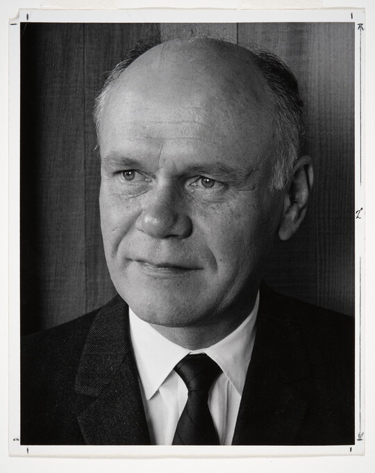 A black-and-white portrait photograph of a mostly bald White man gazing to the side wearing a dark jacket, dark tie, and white shirt.