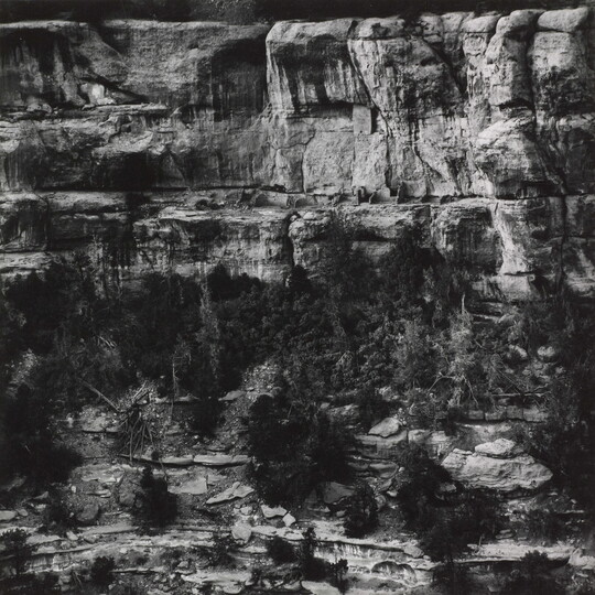 A black-and-white photograph of cliff dwellings in ruins on the side of a steep cliff.