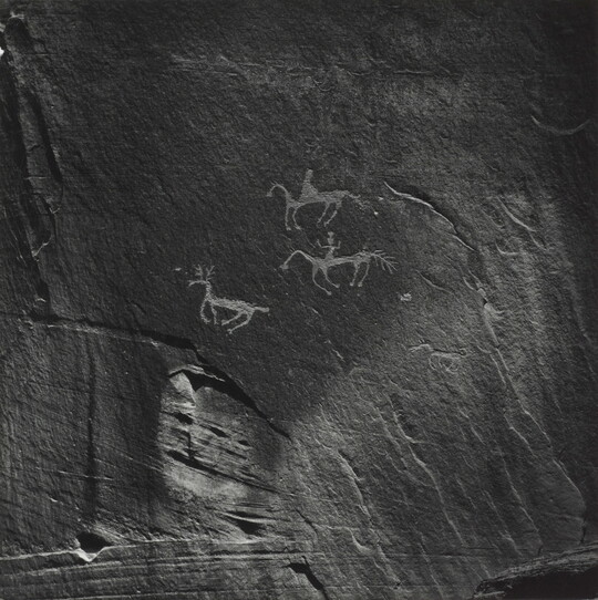 A black-and-white photograph of rock paintings depicting people on horseback and a deer.
