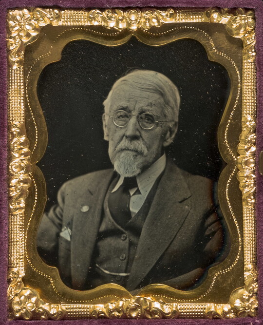 A black-and-white portrait photograph in a decorative gold frame of an older, White man in a suit and glasses.