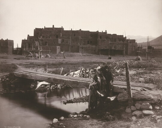 A black-and-white photograph of two Indigenous people by a footbridge over a creek with a large adobe pueblo in the background.