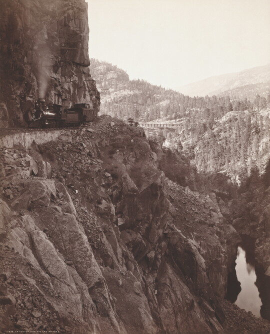 A sepia-toned photograph of a train travelling on the edge of a rocky, mountainside cliff.