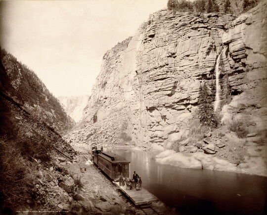 A black-and-white photograph of people on a train at the bottom of a canyon looking at a waterfall across a river.