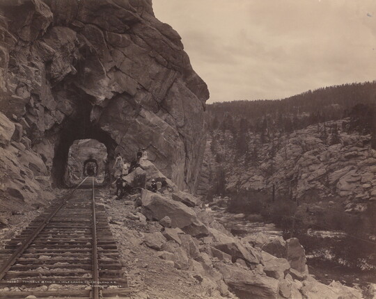 A black-and-white photograph of a train track stretching through a series of rock tunnels in the side of a mountain.