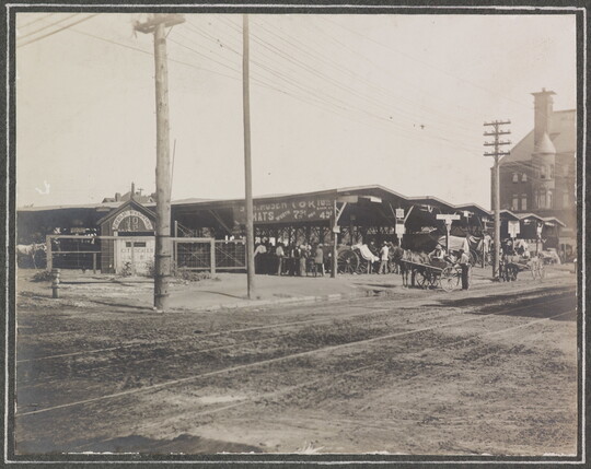 A black-and-white photograph of people and animals under several open-air pavilions next to a dirt road on which horses pull carriages.