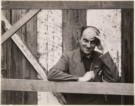 A black-and-white photograph of a bald, White man standing next to a wood structure, wearing a suit jacket and resting his head in his hand.