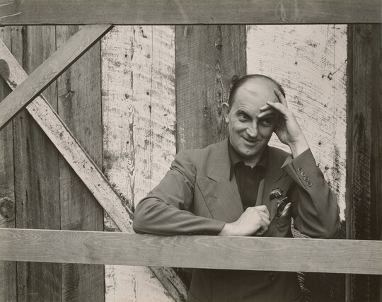 A black-and-white photograph of a bald, White man standing next to a wood structure, wearing a suit jacket and resting his head in his hand.