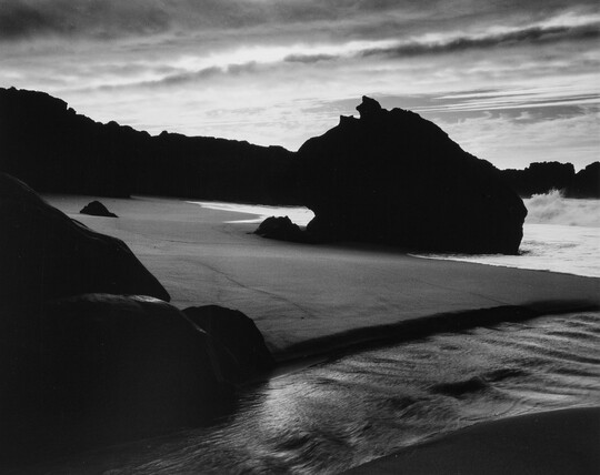 A black-and-white photograph of the sand and sea next to a shore covered with large rocks.