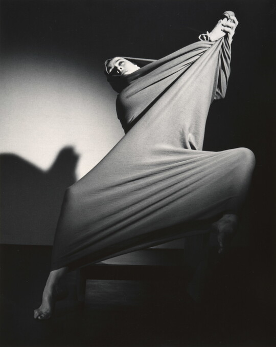 A black-and-white photograph of a modern dancer wrapped in a piece of cloth that she is stretching into shapes over her body.