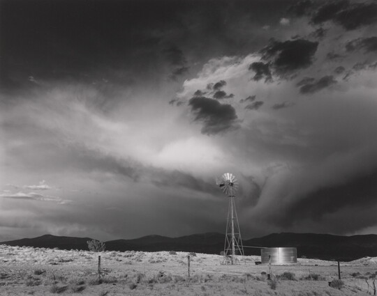 A black-and-white photograph of a windmill, black hills in the distance, under a stormy sky.