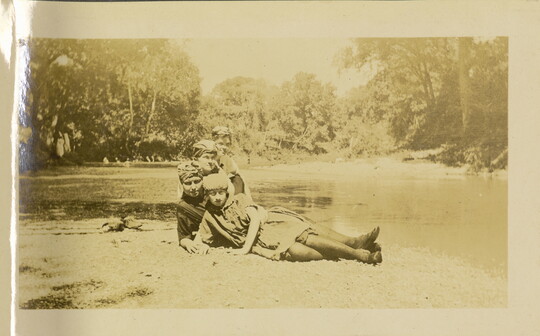 A faded sepia-toned photograph of four White women, all wearing headwraps, posing on the ground next to a river.