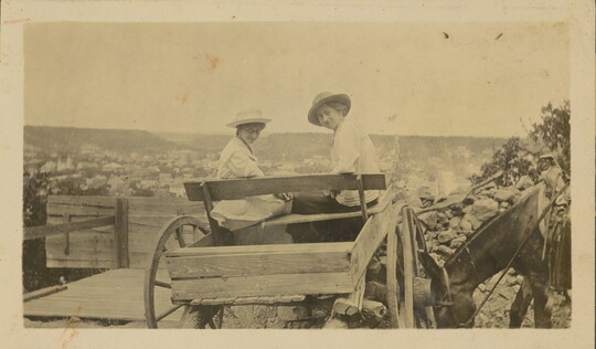 A sepia-toned photograph of two White women wearing sunhats sitting in a cart turned back to face the camera.