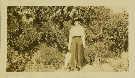 A sepia-toned photograph of a White woman wearing a sunhat and a long skirt, holding a closed parasol and standing near trees and underbrush.