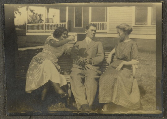 A sepia-toned photograph of two White women and a White man seated between them on a bench in front of a large house.