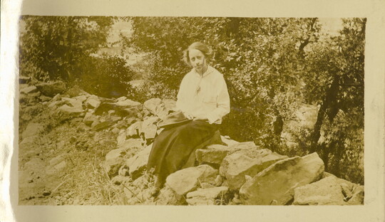 A sepia-toned photograph of a White woman wearing a light blouse and long dark skirt sitting on rocks holding a canvas hat in her lap.