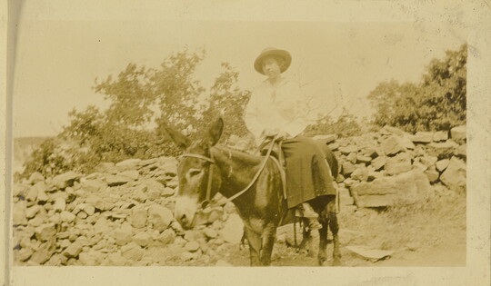 A faded sepia-toned photograph of a White woman wearing a hat, blouse, and skirt, riding a donkey sidesaddle with rocks and trees in the background.
