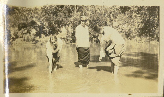 A sepia-toned photograph of three White women standing in calm water holding up their skirts and dipping their hands in the water.