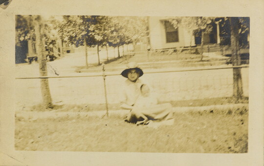 A faded sepia-toned photograph of a White woman in a sunhat holding a baby wearing a bonnet on her lap sitting on grass in front of a fence.