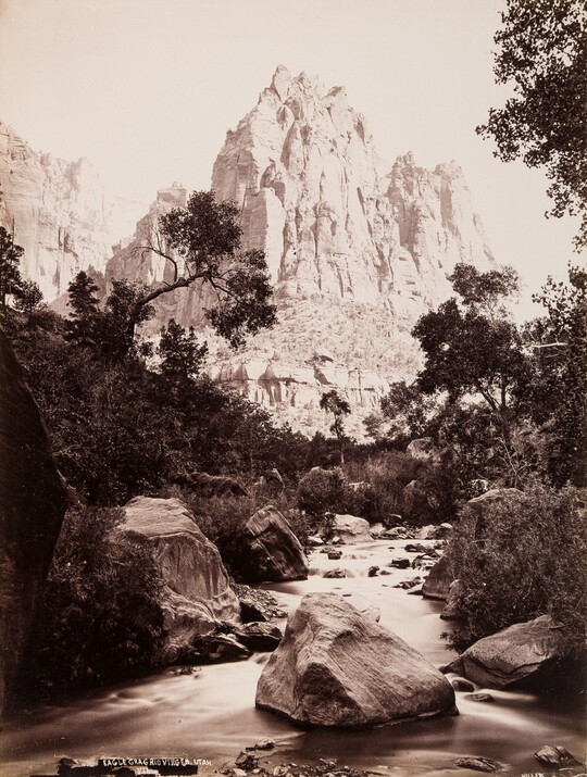 A black-and-white photograph of a stream surrounded by boulders and vegetation and a rugged cliff in the background.