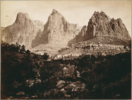 A black-and-white photograph of three large, majestic crags with rocks and vegetation in the foreground.