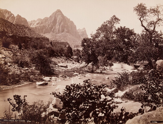A black-and-white photograph of a rocky river surrounded by vegetation, and a tall mountain rising in the background.