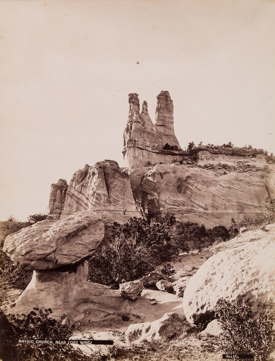A sepia-toned photograph of a tall rock formation on top of large boulders above scrubby vegetation.