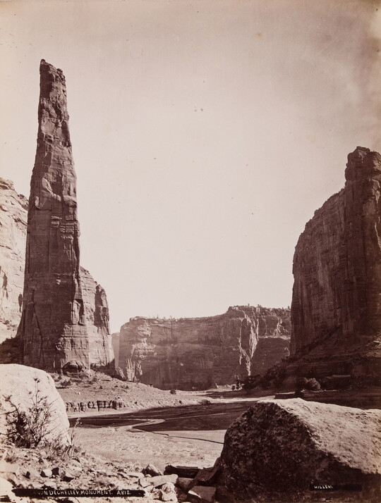 A sepia-toned photograph of a tall, narrow rock formation opposite a butte with a horizontal rock formation in the background.