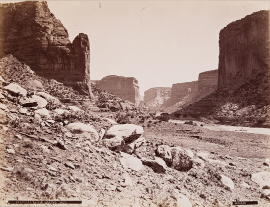 A sepia-toned photograph of buttes and other rock formations rising above a narrow canyon covered with loose rocks.