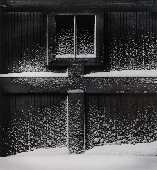 A black-and-white photograph of a garage door with snow building up on the wood beams and a small window.