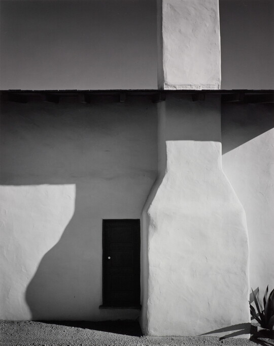 A black-and-white photograph of the side of an adobe house with the foundation of a chimney, shadows on the wall, and a door.