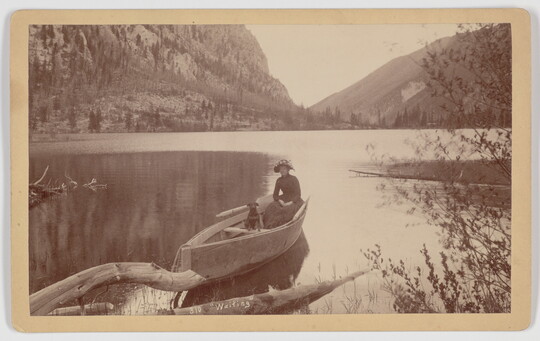 A sepia-toned photograph of a woman and a dog in a canoe on a still lake in the mountains.