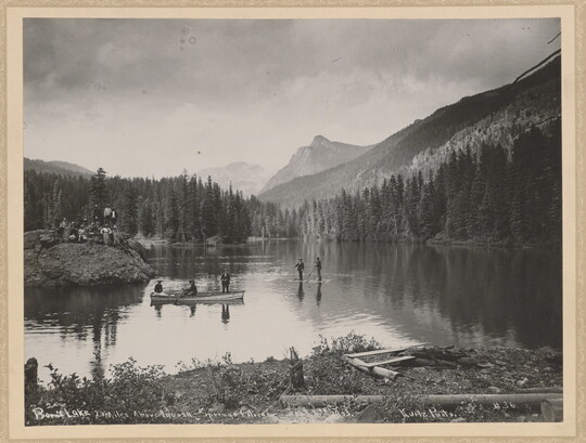 A black-and-white photograph of people canoeing and fishing on a lake surrounded by trees and mountains.