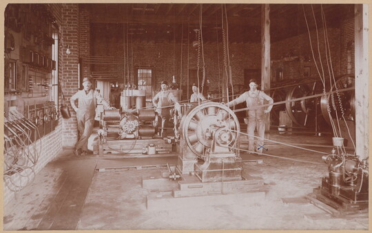 A sepia-toned photograph of several White men posing by industrial equipment and gears in a brick building.