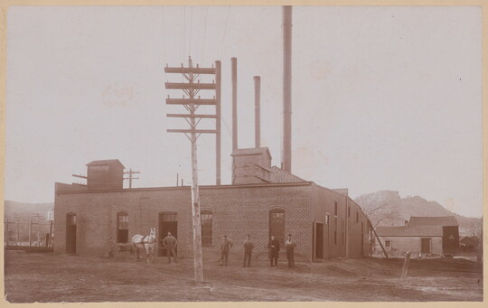 A sepia-toned photograph of men standing in front of a one-story brick building with smokestacks.