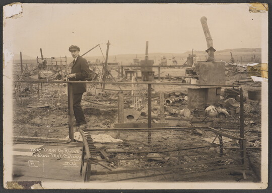 A black-and-white photograph of a White man standing among the ruins of a village.