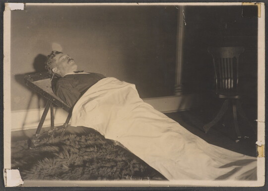 A black-and-white photograph of a White man in a formal suit laying on a stretcher and partially covered by a white sheet.