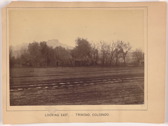 A sepia-toned photograph of train tracks in the foreground, a homestead in the mid-ground, and mountains in the background.