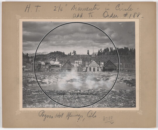 A black-and-white photograph of a hot spring with steam rising off the water and houses and hills in the background.