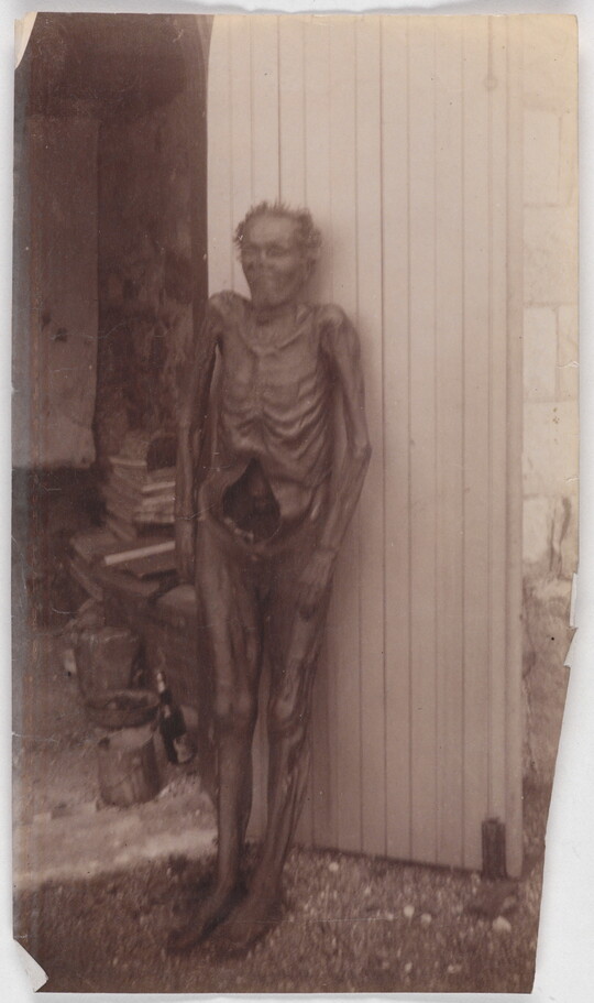 A black-and-white photograph of a mummified person standing upright and leaning against a wall.