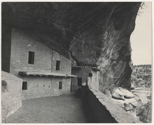A black-and-white photograph of a man standing in a stone cliff dwelling overlooking the valley beyond.