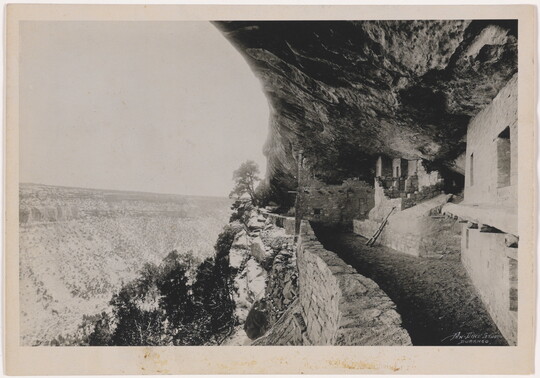 A black-and-white photograph of human-made structures built into a steep cliff face overlooking a deep valley.