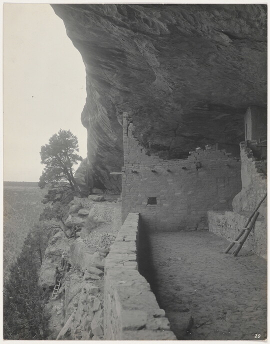 A black-and-white photograph of a dirt path next to human-made structures built under a cliff overhang.