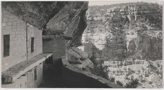 A black-and-white photograph of human-made stone structures in a cliff face overlooking a cliff wall across a valley.
