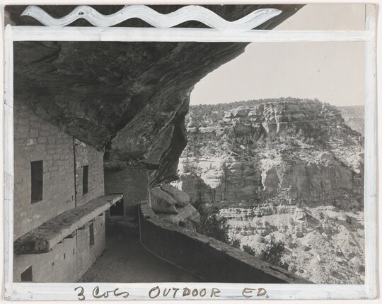 A black-and-white photograph of human-made stone structures in a cliff face overlooking a cliff wall across a valley; white paint marks how it should be cropped.