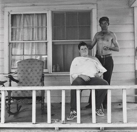 A black-and-white photograph of two White men on the porch of a house: one seated with a white apron around his neck and shoulders; the other standing, shirtless, holding clippers in one hand, the other on the seated man's head.