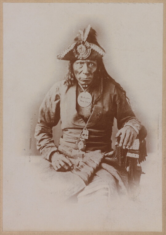 A black-and-white portrait photograph of a seated Indigenous man seated wearing a colonial-style jacket and a tricorn hat.