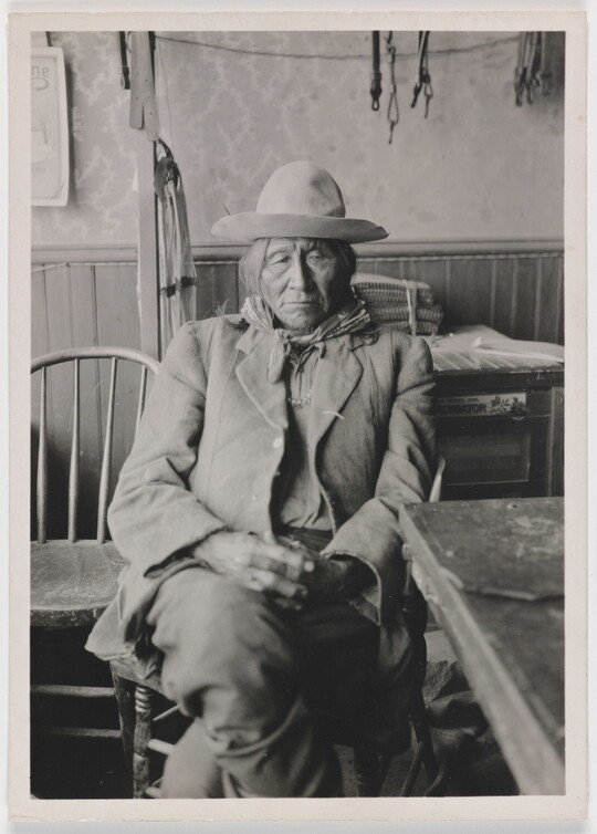A black-and-white photograph of an Indigenous man in a rumpled suit seated next to a table with his hands in his lap.