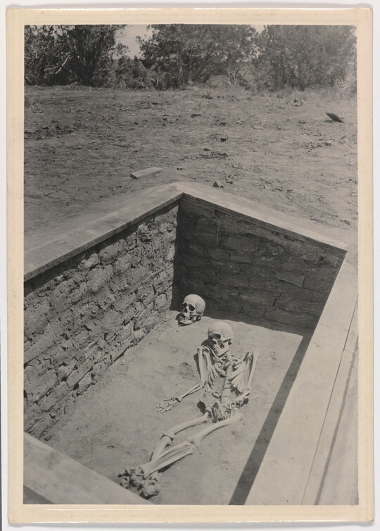 A black-and-white photograph of an archaeological grave site with a full human skeleton laying beside a skull.
