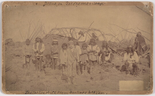 A slightly faded sepia-toned photograph of a group of Indigenous adults and children and one White child standing in front of a camp.