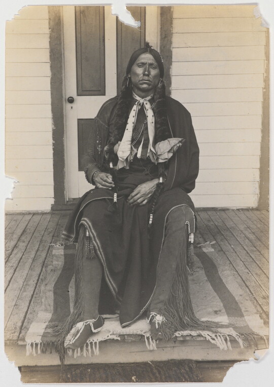 A black-and-white photograph of an Indigenous man with long braids seated on a wooden porch.